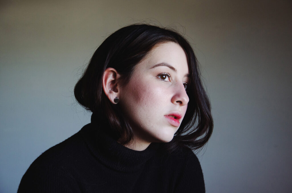 Gabriela Inzunza a thoughtful individual gazes into the distance her dark hair cascading down her shoulder and her ear adorned with a silver earring.